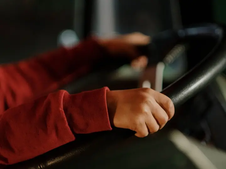 Hands in red sleeves resting on a large steering wheel inside a dim cab