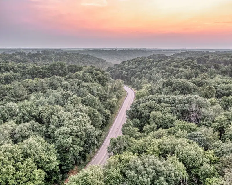 Aerial view of a road curving through wooded hills under a pink evening sky